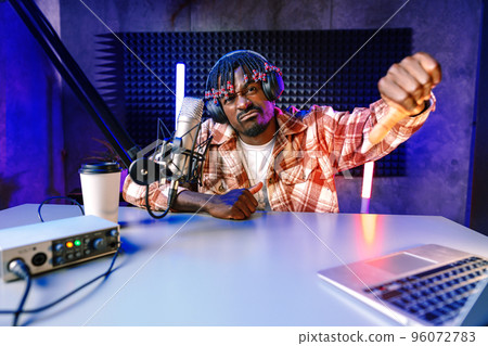 African radio host sitting at desk recording in studio with microphone and laptop 96072783
