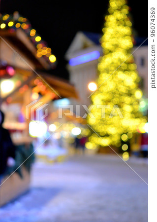Blurred background. Christmas New Year tree decorated yellow garland illumination, small wooden houses garlands of lanterns on roof and silhouettes of people walking along city street. 96074369