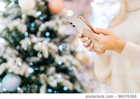 Close-up cropped shot of unrecognizable young woman using typing smartphone standing in hall of celebrate shopping mall in Christmas eve, on background of bright beautiful xmas decorations. Close-up cropped shot of unrecognizable young woman using typing smartphone standing in hall of celebrate shopping mall in Christmas eve, on background of bright beautiful xmas decorations. 96076368