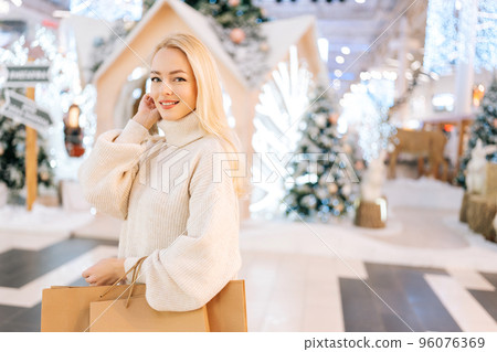 Portrait of charming young woman with blonde hair holding bags with purchase smiling looking at camera standing in hall of celebrate shopping mall in Christmas eve, on background of xmas decorations. Portrait of charming young woman with blonde hair holding bags with purchase smiling looking at camera standing in hall of celebrate shopping mall in Christmas eve, on background of xmas decorations. 96076369