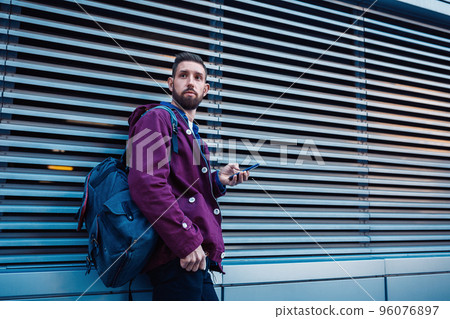 Confident bearded man holding in and mobile phone while standing near street wall in sunny summer evening. 96076897