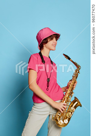 Portrait of cheerful teen girl with curly brown short hair posing in pink panama and playing saxophone isolated over blue background 96077470