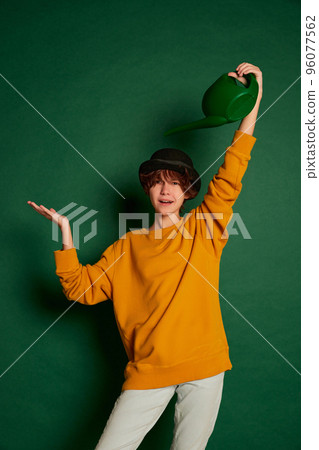 Portrait of cheerful teen girl with curly brown short hair posing in yellow sweater and hat isolated over green background. Watering head 96077562