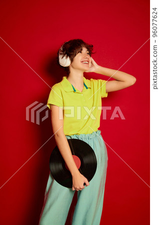 Portrait of cheerful teen girl with curly brown short hair listening to music in headphones isolated over red background. Vinyl 96077624