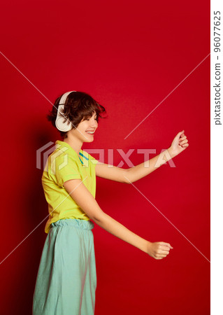 Portrait of cheerful teen girl with curly brown short hair posing, listening o music in headphones and dancing isolated over red background 96077625