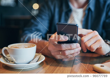 Man hands in denim shirt slide with finger on screen his smart phone, near cup with coffee on wooden table Man hands in denim shirt slide with finger on screen his smart phone, near cup with coffee on wooden table 96077789