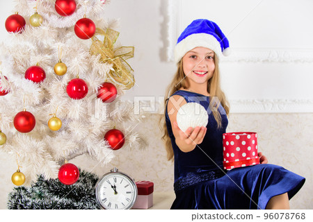Happy new year concept. Kid girl sit near christmas tree hold gift box. Little girl santa hat excited about christmas gift or present. Child celebrate christmas at home. Time to open christmas gifts 96078768