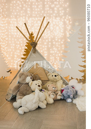 Children's photo zone in the Christmas photo studio - wigwam with teddy bears on the background of silhouettes of fir trees and garlands. Children's photo zone in the Christmas photo studio - wigwam with teddy bears on the background of silhouettes of fir trees and garlands. 96080710