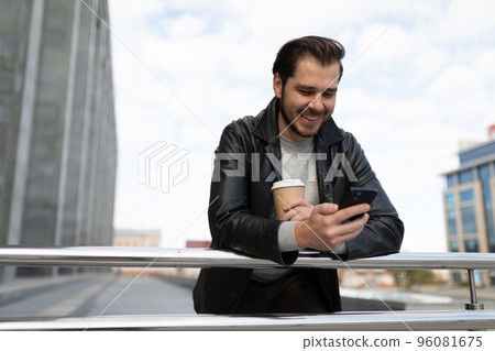 an adult man outside with a cup of coffee in his hands looks at the screen of a mobile phone 96081675