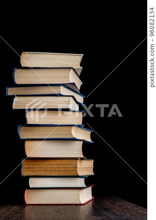 Tall stack of hardback books on a wooden surface against a dark background. 96082134