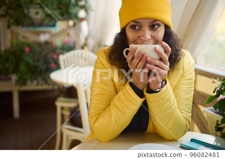 Close up portrait of millennial woman in bright yellow hat and jacket, warming her hands with a cup of hot drink, taking a sip of fragrant coffee, relaxing and enjoying a day off in a cozy coffee shop Close up portrait of millennial woman in bright yellow hat and jacket, warming her hands with a cup of hot drink, taking a sip of fragrant coffee, relaxing and enjoying a day off in a cozy coffee shop 96082481