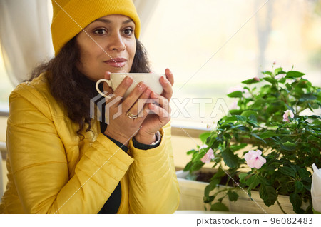 Close-up portrait of a millennial brunette in bright yellow hat and jacket, warming her hands with a cup of hot drink, sitting at table by a window overlooking a beautiful autumn park in a coffee shop 96082483
