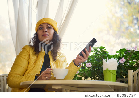 Delighted curly brunette pretty woman in yellow jacket and hat, with smartphone in her hands, looking thoughtfully aside while enjoying her coffee break in a cozy cafe on a sunny warm autumn day Delighted curly brunette pretty woman in yellow jacket and hat, with smartphone in her hands, looking thoughtfully aside while enjoying her coffee break in a cozy cafe on a sunny warm autumn day 96083757