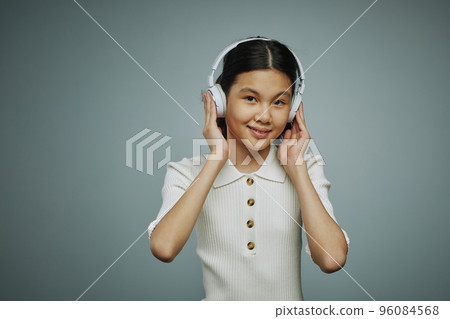 Smiling youthful Asian girl touching headphones while listening to music Smiling youthful Asian girl touching headphones while listening to music 96084568