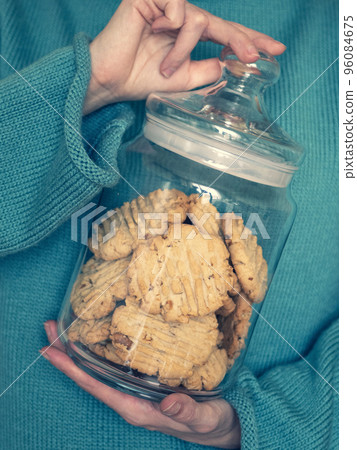 Young woman in warm turquois sweater is keeping the glass jar with homemade nut cookies in her hands Young woman in warm turquois sweater is keeping the glass jar with homemade nut cookies in her hands 96084675