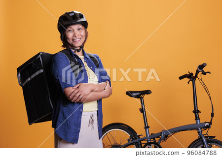 Asian deliver carrying food takeaway backpack standing beside bike in studio with yellow background. Restaurant courier delivering pizza for dinner. Food transportation service and takeaway concept 96084788