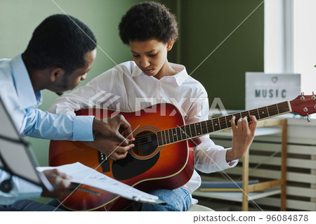 Youthful diligent girl playing acoustic guitar while teacher consulting her 96084878