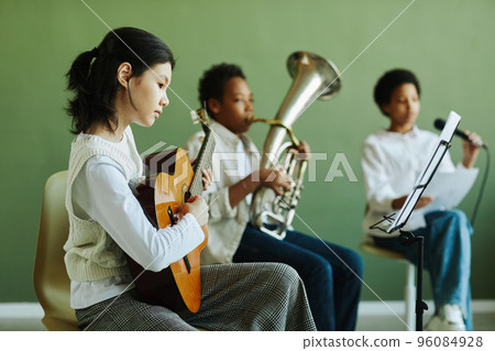 Cute Asian schoolgirl playing acoustic guitar and looking at notes on stand Cute Asian schoolgirl playing acoustic guitar and looking at notes on stand 96084928
