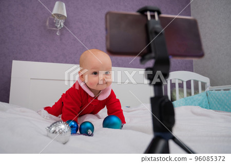 A baby boy dressed in a Santa suit sits on the bed and looks at the sick phone. Christmas greetings by video call A baby boy dressed in a Santa suit sits on the bed and looks at the sick phone. Christmas greetings by video call 96085372