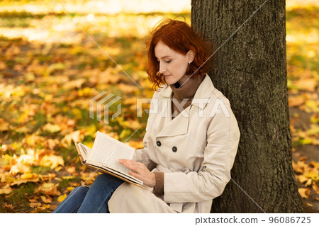 Concentrated smart caucasian millennial female with red hair in raincoat reads book and thinks 96086725