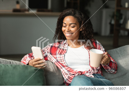 Cheerful black young woman with cup of drink, chatting on phone, reading blog, enjoy comfort Cheerful black young woman with cup of drink, chatting on phone, reading blog, enjoy comfort 96086807