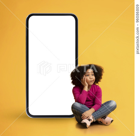 Pensive Black Little Girl Sitting Next To Big Blank Smartphone Pensive Black Little Girl Sitting Next To Big Blank Smartphone 96086809