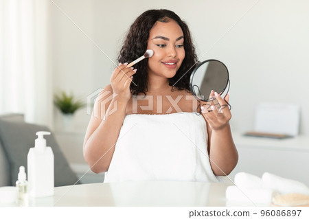 Beautiful african american chubby woman applying decorative makeup, holding mirror, making daily beauty routine at home Beautiful african american chubby woman applying decorative makeup, holding mirror, making daily beauty routine at home 96086897