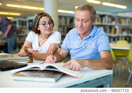 Young female Latina tutor teaching a mature caucasian elder Young female Latina tutor teaching a mature caucasian elder 96087728