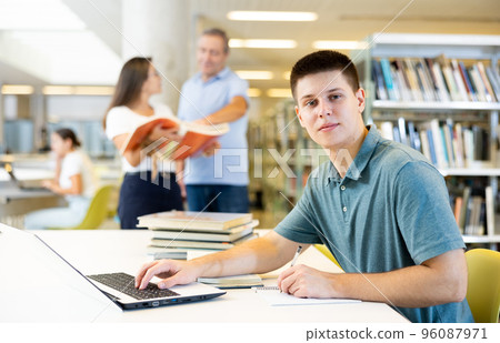 Focused casual young male writing in planner and using laptop in the library 96087971