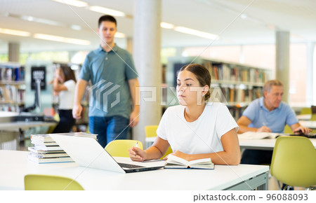 Focused European casual female writing in notepad and using laptop in the library Focused European casual female writing in notepad and using laptop in the library 96088093