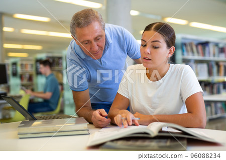 Mature male explaining information for casual female while she is reading books in the library 96088234