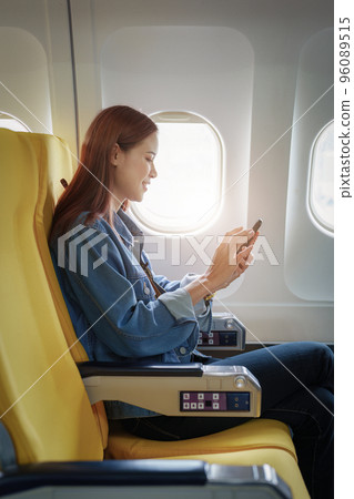 Attractive portrait of Asian woman sitting at window seat in economy class using mobile phone during plane flight, travel concept, vacation. Attractive portrait of Asian woman sitting at window seat in economy class using mobile phone during plane flight, travel concept, vacation. 96089515