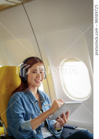 Attractive portrait of an Asian woman sitting in a window seat in economy class using tablet and listening to instrumental music during an airplane flight, travel concept, vacation, relaxation. 96089725