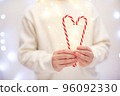 Child in white sweater holding a heart-shaped lollipop cane on light bokeh background. 96092330
