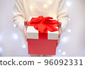 Child in white sweater holding red Christmas gift in hand on light bokeh background 96092331