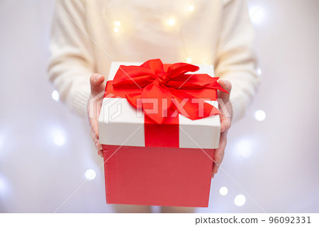 Child in white sweater holding red Christmas gift in hand on light bokeh background 96092331