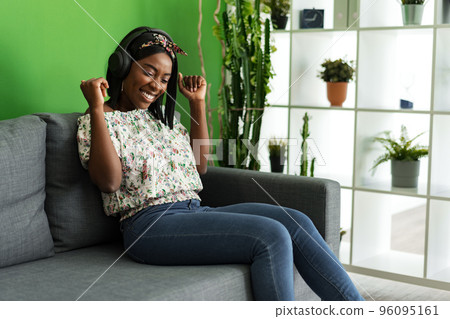 African woman listening to music with headphones while sitting on couch at home 96095161