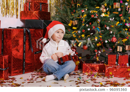 Caucasian boy and sits on floor with gifts near Christmas tree. Child in red 96095360