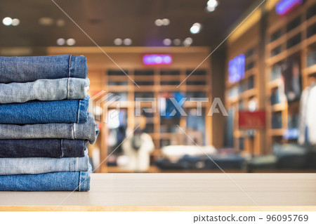 Blue denim jeans stack on wood table top with abstract blur clothing store in modern shopping mall defocused bokeh light background Blue denim jeans stack on wood table top with abstract blur clothing store in modern shopping mall defocused bokeh light background 96095769