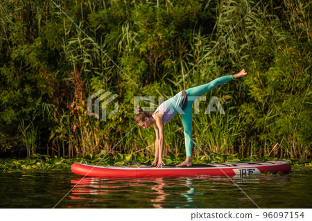 Photo of young woman doing hand stand on stand up paddle board. She wearing a leggings and top. 96097154