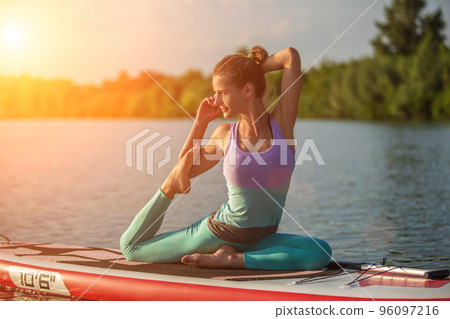 Young woman sitting on paddle board, practicing yoga pose. Doing yoga exercise on sup board, active summer rest 96097216