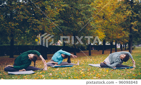 Good-looking sportswomen are doing stretching exercises sitting on yoga mats in city park at autumn weekend. Leisure activity, common hobby and youth concept. Good-looking sportswomen are doing stretching exercises sitting on yoga mats in city park at autumn weekend. Leisure activity, common hobby and youth concept. 96098848