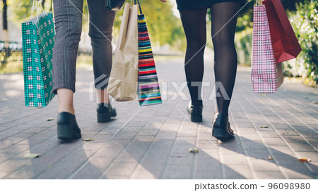 Low angle shot of women's legs walking in the street with colorful paper bags after busy day in shops. Young people, leisure, shopping and friendship conccept. Low angle shot of women's legs walking in the street with colorful paper bags after busy day in shops. Young people, leisure, shopping and friendship conccept. 96098980