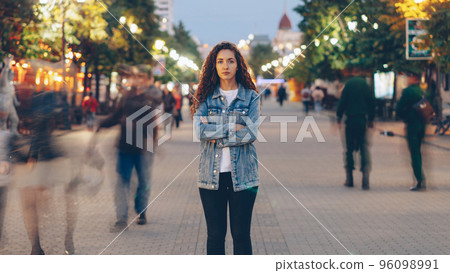 Portrait of stylish young lady tired of usual haste standing in the street among whizzing people and looking at camera. Time, youth and modern society concept. Portrait of stylish young lady tired of usual haste standing in the street among whizzing people and looking at camera. Time, youth and modern society concept. 96098991