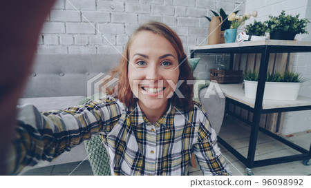 Portrait of happy young woman looking at camera, holding gadget and talking to friends online smiling and gesturing standing at home. Communication, technology and people concept. Portrait of happy young woman looking at camera, holding gadget and talking to friends online smiling and gesturing standing at home. Communication, technology and people concept. 96098992