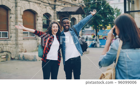 Young woman is taking pictures of beautiful couple African American guy and Caucasian girl tourists standing in the street and posing for camera. Tourism, technology and friends concept. 96099006