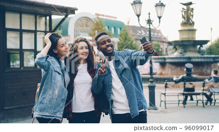 Happy African American guy is taking selfie with friends beautiful Caucasian girls standing in street posing and holding modern smartphone during journey in foreign country. Happy African American guy is taking selfie with friends beautiful Caucasian girls standing in street posing and holding modern smartphone during journey in foreign country. 96099017