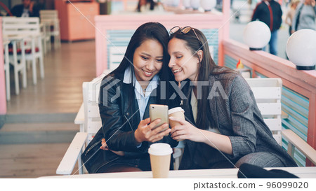 Pretty young ladies friends are using modern smartphone sitting at table in cafe with take away coffee. Happy girls are touching screen, watching funny photos and laughing. Pretty young ladies friends are using modern smartphone sitting at table in cafe with take away coffee. Happy girls are touching screen, watching funny photos and laughing. 96099020