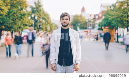 portrait of serious young man looking at camera standing in center of busy pedestrian street in summer wearing stylish clothing while many people are passing by. portrait of serious young man looking at camera standing in center of busy pedestrian street in summer wearing stylish clothing while many people are passing by. 96099039