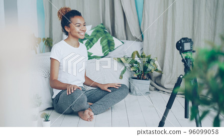 Pretty African American teenager vlogger is recording video for social media followers, girl is looking at camera on tripod, speaking and gesturing sitting on bedroom floor. Pretty African American teenager vlogger is recording video for social media followers, girl is looking at camera on tripod, speaking and gesturing sitting on bedroom floor. 96099058
